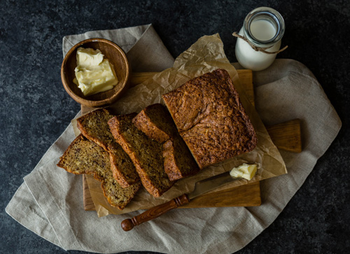 Sliced loaf of banana bread on cutting board with butter and milk